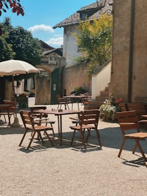 A quaint outdoor caf&eacute; setting with several wooden tables and chairs arranged on a gravel surface. A large white umbrella provides shade for one of the tables. The area is enclosed by rustic buildings with textured walls and a staircase leading up to a higher entrance. Lush greenery and potted plants add a touch of nature, with a clear blue sky and fluffy clouds visible overhead.