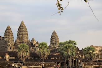 A historic temple with intricately carved stone towers stands majestically against a partly cloudy sky. In the foreground, a monkey sits atop the temple structure, while several palm trees add greenery to the scene.