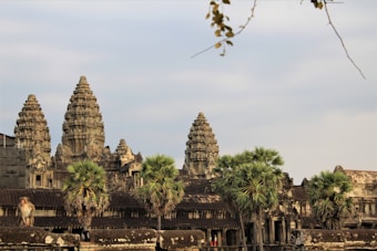 A historic temple with intricately carved stone towers stands majestically against a partly cloudy sky. In the foreground, a monkey sits atop the temple structure, while several palm trees add greenery to the scene.