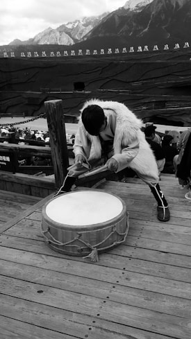 A person wearing traditional clothing and fur appears to be playing a large drum on a wooden platform. In the background, there is a crowd seated in an amphitheater setting, with a mountainous landscape and a line of people standing in the distance on a raised platform.