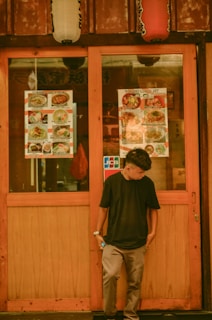 A person stands in front of a restaurant entrance displaying two menu posters, featuring a variety of dishes. Above the entrance, traditional lanterns add to the cultural ambiance. The person's casual attire suggests a relaxed vibe.