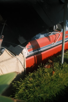 A close-up view of a red vintage vehicle partially covered by a protective tarp, with the badge 'F100 Custom' visible. The scene includes lush green plants in the foreground, with sunlight casting shadows on both the car and the greenery.