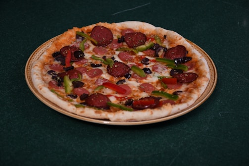 A pizza with various toppings including slices of pepperoni, green bell peppers, black olives, and red bell peppers is placed on a plate. The crust appears lightly baked with a golden brown edge, and the cheese is melted evenly across the surface.