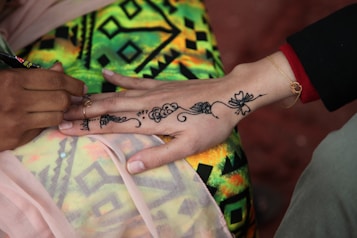 Henna is being applied to a person's hand with intricate floral and geometric patterns. The hand is resting against a colorful, patterned fabric, with hints of geometric designs. The scene captures the tattooing process with focus on the details of the artwork.