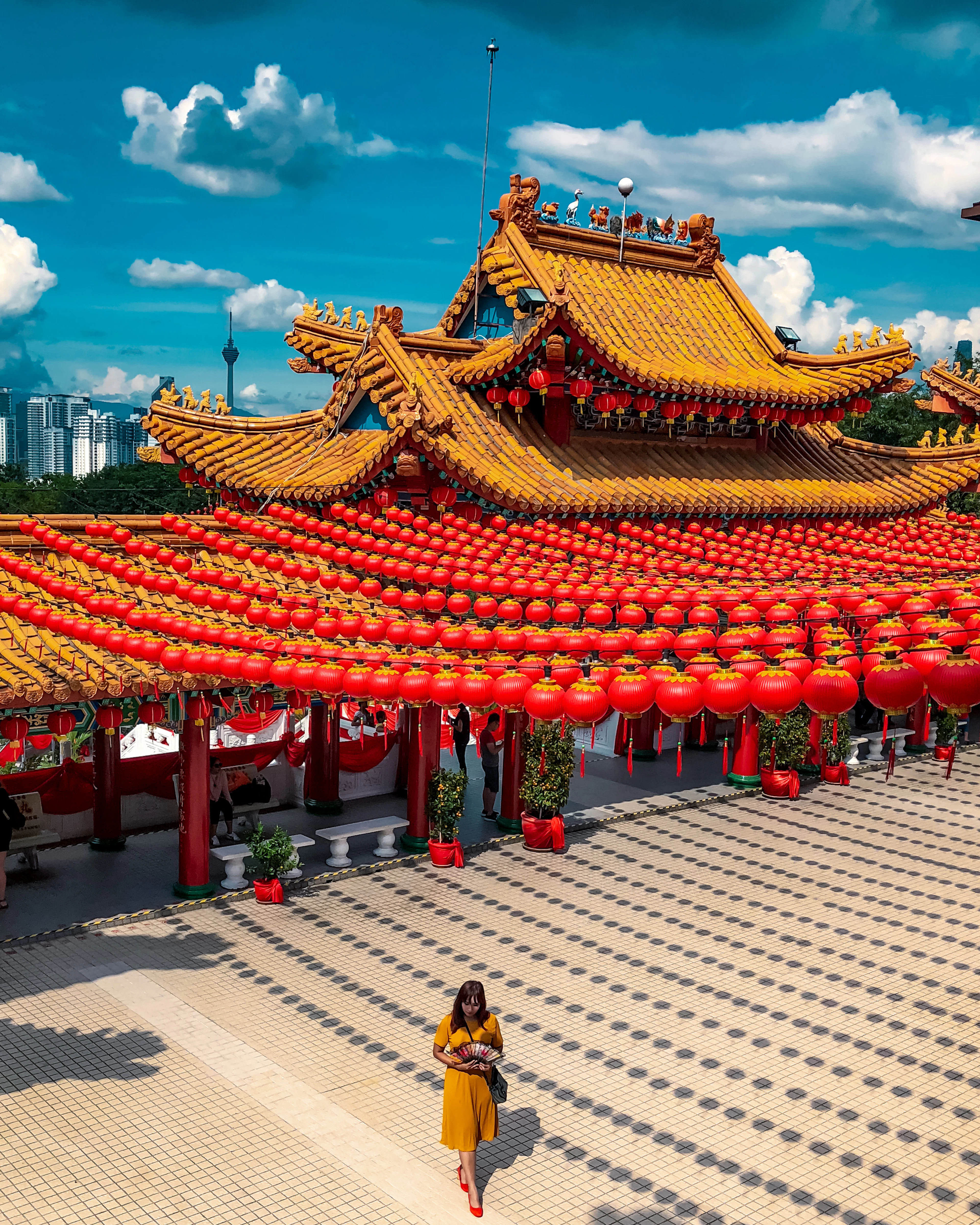 Thousands of red lanterns strung in long rows beneath the tiered gold and vermilion roof of Thean Hou Temple in Kuala Lumpur, with the KL Tower and city towers on the skyline behind.