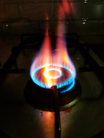 A close-up view of a lit gas stove burner with visible blue and orange flames. The stove grate is discernible in the background, casting shadowed lines on the stovetop surface.