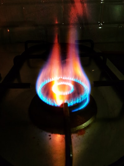 A close-up view of a lit gas stove burner with visible blue and orange flames. The stove grate is discernible in the background, casting shadowed lines on the stovetop surface.