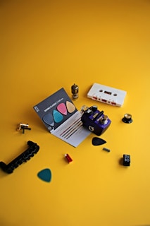 A variety of small, colorful items are spread across a yellow surface. These include guitar picks in different colors, a small electronic component resembling a vacuum tube, a white cassette tape, a purple effects pedal, and some other small mechanical parts. A card displaying descriptions of the guitar picks is also present.