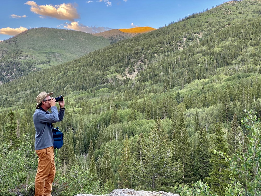 man in blue jacket and brown pants carrying black backpack standing on rock near green trees