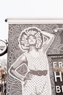 A large mural on the side of a building features a woman with an afro, wearing a polka dot outfit. Wrapped around her hair are the words 'follow your dreams'. The background consists of intricate, patterned designs. Nearby, part of a building with glass windows is visible, along with a small tree and part of a street.