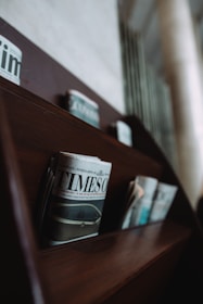 A wooden shelf holds several newspapers, including a prominently displayed one titled 'TIMES'. The newspapers are arranged in neat rows, and the shelf is part of a dimly lit interior with a blurred background.