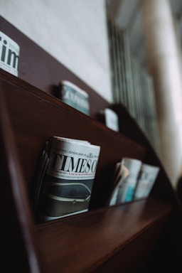 A wooden shelf holds several newspapers, including a prominently displayed one titled 'TIMES'. The newspapers are arranged in neat rows, and the shelf is part of a dimly lit interior with a blurred background.