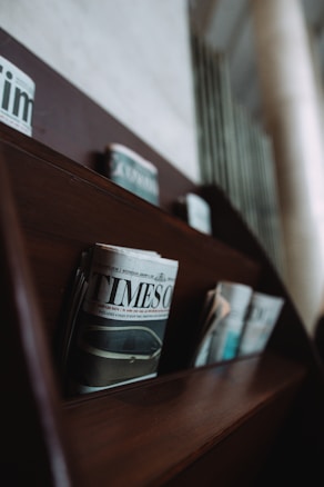 A wooden shelf holds several newspapers, including a prominently displayed one titled 'TIMES'. The newspapers are arranged in neat rows, and the shelf is part of a dimly lit interior with a blurred background.