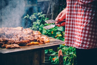 A person wearing a red and white checkered apron holds a pair of spatulas while grilling a variety of meat pieces on a barbecue. Smoke is rising from the grill and there is lush greenery in the background.