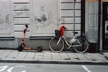 A red electric scooter and a white bicycle are parked side by side on a sidewalk against a beige-colored wall with peeling paint and grime. The bicycle has a black basket in the front with a red, fluffy toy or object in it. A black and white helmet is placed on the bicycle's frame.