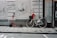 A red electric scooter and a white bicycle are parked side by side on a sidewalk against a beige-colored wall with peeling paint and grime. The bicycle has a black basket in the front with a red, fluffy toy or object in it. A black and white helmet is placed on the bicycle's frame.