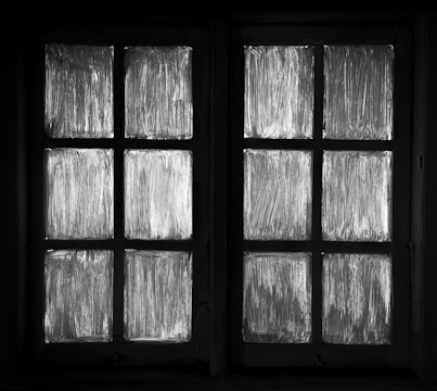 A black and white image featuring a window with four panes on each side. The window panes are covered in streaked, semi-transparent material, giving a frosted appearance.