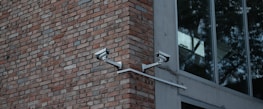 Two security cameras mounted on a red brick wall are angled towards each other. Adjacent to the brick wall is a modern building with large glass windows reflecting trees.