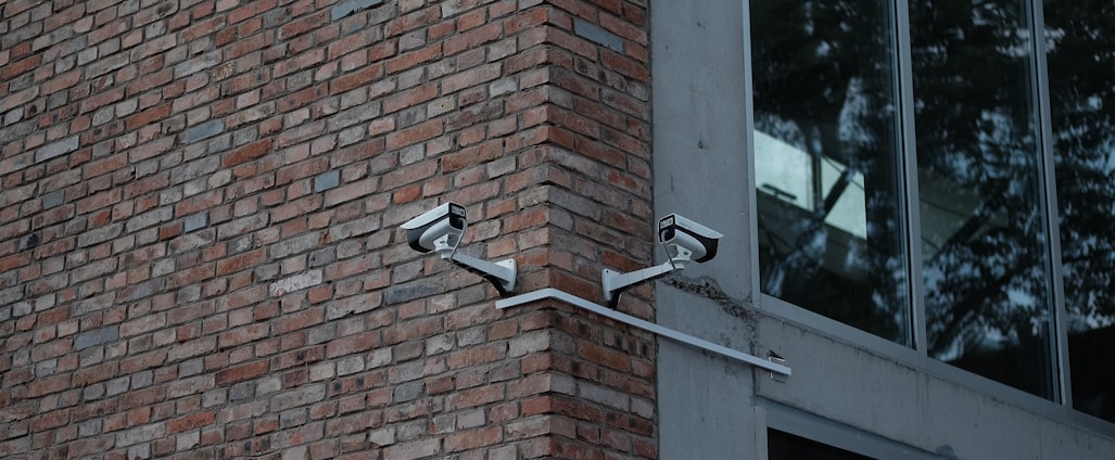 Two security cameras mounted on a red brick wall are angled towards each other. Adjacent to the brick wall is a modern building with large glass windows reflecting trees.