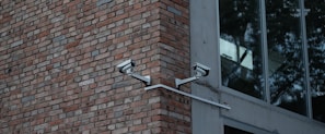 Two security cameras mounted on a red brick wall are angled towards each other. Adjacent to the brick wall is a modern building with large glass windows reflecting trees.