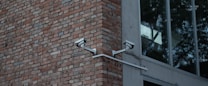Two security cameras mounted on a red brick wall are angled towards each other. Adjacent to the brick wall is a modern building with large glass windows reflecting trees.