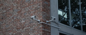 Two security cameras mounted on a red brick wall are angled towards each other. Adjacent to the brick wall is a modern building with large glass windows reflecting trees.