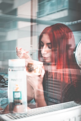 A woman with red hair is seated indoors, holding a jar of Pantene hair product in her hand. She appears to be gazing thoughtfully at the jar, with a window reflection showing parts of the surroundings. There is another bottle of Pantene product on the table in front of her.