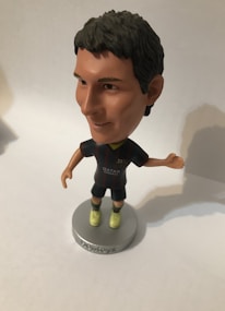 A bobblehead figure of a soccer player wearing a dark jersey with a logo and 'Qatar Airways' printed on it. The figure has short dark hair, light-colored shoes, and is mounted on a round base with the word 'soccerstarz'. The background is neutral, likely a plain surface.