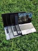 Multiple laptops are stacked on top of each other, placed on a grassy surface. The screen of the topmost laptop displays a reflection of a building and a railing.