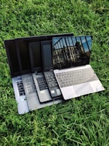 Multiple laptops are stacked on top of each other, placed on a grassy surface. The screen of the topmost laptop displays a reflection of a building and a railing.