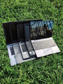 Multiple laptops are stacked on top of each other, placed on a grassy surface. The screen of the topmost laptop displays a reflection of a building and a railing.