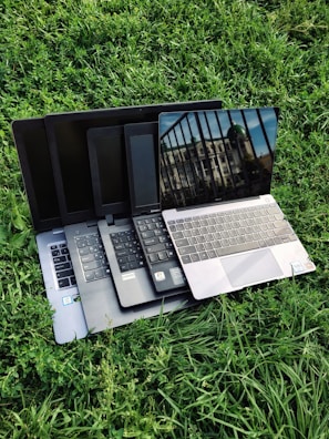 Multiple laptops are stacked on top of each other, placed on a grassy surface. The screen of the topmost laptop displays a reflection of a building and a railing.