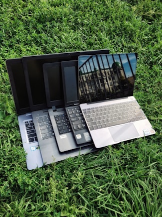 Multiple laptops are stacked on top of each other, placed on a grassy surface. The screen of the topmost laptop displays a reflection of a building and a railing.