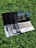 Multiple laptops are stacked on top of each other, placed on a grassy surface. The screen of the topmost laptop displays a reflection of a building and a railing.