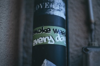A close-up photograph of a dark cylindrical object, possibly a pipe or pole, featuring a sticker with the words 'smoke weed every day' in stylized, bold letters. The background includes part of a textured wall and some metal fixtures.