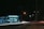 A gas station, illuminated by artificial lighting, operates at night with visible signage reading 'Posto Meridional'. The forecourt is quiet with minimal activity. Street lights illuminate the surrounding road, capturing the light trails of passing vehicles, giving a sense of motion and energy.