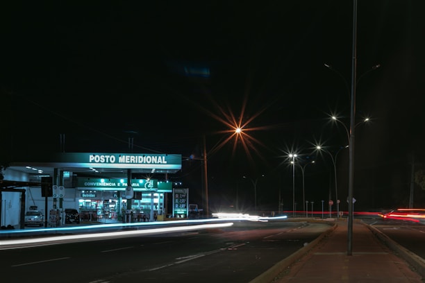 A gas station, illuminated by artificial lighting, operates at night with visible signage reading 'Posto Meridional'. The forecourt is quiet with minimal activity. Street lights illuminate the surrounding road, capturing the light trails of passing vehicles, giving a sense of motion and energy.