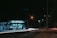 A gas station, illuminated by artificial lighting, operates at night with visible signage reading 'Posto Meridional'. The forecourt is quiet with minimal activity. Street lights illuminate the surrounding road, capturing the light trails of passing vehicles, giving a sense of motion and energy.