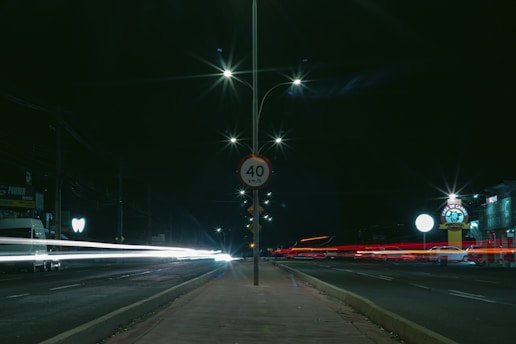 A nighttime urban scene with long exposure light trails from passing vehicles on either side of a central road. A speed limit sign indicating 40 km/h is prominently displayed on a lamppost. Streetlights cast bright flares in the dark sky, illuminating the otherwise dim surroundings. Buildings and signs are visible in the background, with some glowing due to artificial lighting.