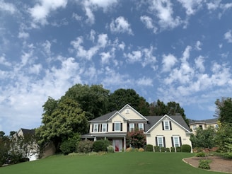 A large, two-story house with a classic architectural style is surrounded by lush greenery and a well-manicured lawn. The sky above is bright blue with scattered white clouds, creating a serene and inviting atmosphere.