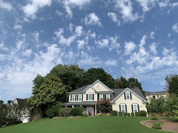 A large, two-story house with a classic architectural style is surrounded by lush greenery and a well-manicured lawn. The sky above is bright blue with scattered white clouds, creating a serene and inviting atmosphere.