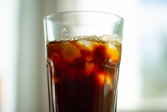 A tall glass filled with a dark, carbonated beverage, featuring ice cubes and a layer of bubbles near the rim. The background is softly blurred, suggesting a bright and airy setting.