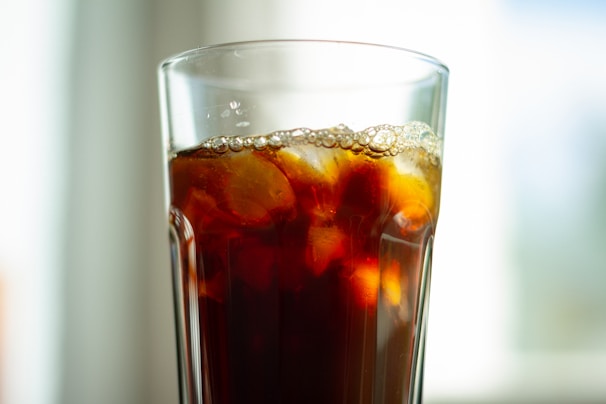 A tall glass filled with a dark, carbonated beverage, featuring ice cubes and a layer of bubbles near the rim. The background is softly blurred, suggesting a bright and airy setting.