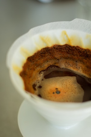 A close-up view of a coffee brewing process with a paper filter in a pour-over setup. The freshly brewed coffee is visible with a layer of crema on top. The filter is slightly stained with coffee grounds, indicating recent use.
