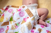 A baby lies on a soft surface wearing a diaper, partially covered by a colorful blanket that features various ice cream and dessert illustrations. The baby is positioned on its side with one leg bent, resting on a light yellow background.