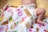 A baby lies on a soft surface wearing a diaper, partially covered by a colorful blanket that features various ice cream and dessert illustrations. The baby is positioned on its side with one leg bent, resting on a light yellow background.