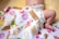 A baby lies on a soft surface wearing a diaper, partially covered by a colorful blanket that features various ice cream and dessert illustrations. The baby is positioned on its side with one leg bent, resting on a light yellow background.