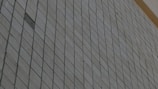 A close-up view of a wall composed of square tiles arranged in a diagonal pattern with a slight reflection. The tiles appear to be light gray or beige, with lines outlining each square tile.