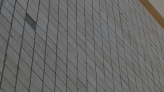 A close-up view of a wall composed of square tiles arranged in a diagonal pattern with a slight reflection. The tiles appear to be light gray or beige, with lines outlining each square tile.