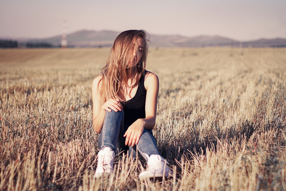 woman in black tank top and blue denim jeans sitting on brown grass field during daytime